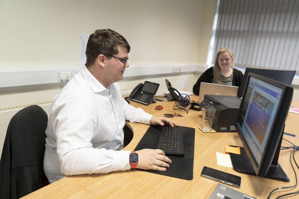 male in a white shirt sat at desk on a computer.
