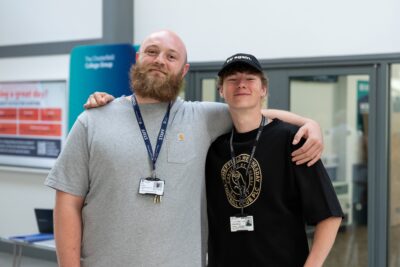 Member of staff in grey t shirt and student in black t shirt posing for a smily photo